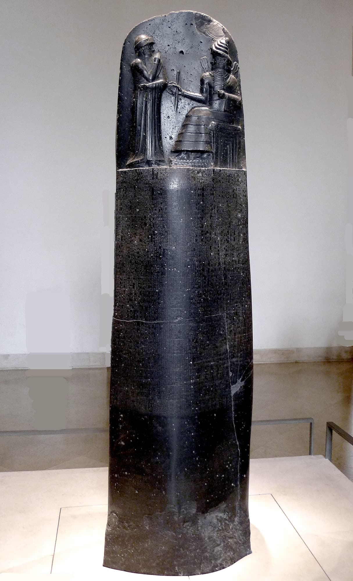 A tall, dark basalt stone pillar in a museum, with a carved scene of two figures at the top and dense rows of cuneiform writing below.