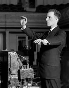 Black-and-white photo of Leon Theremin in a suit, one hand raised beside a tall vertical antenna and the other held flat over the instrument, without touching it.