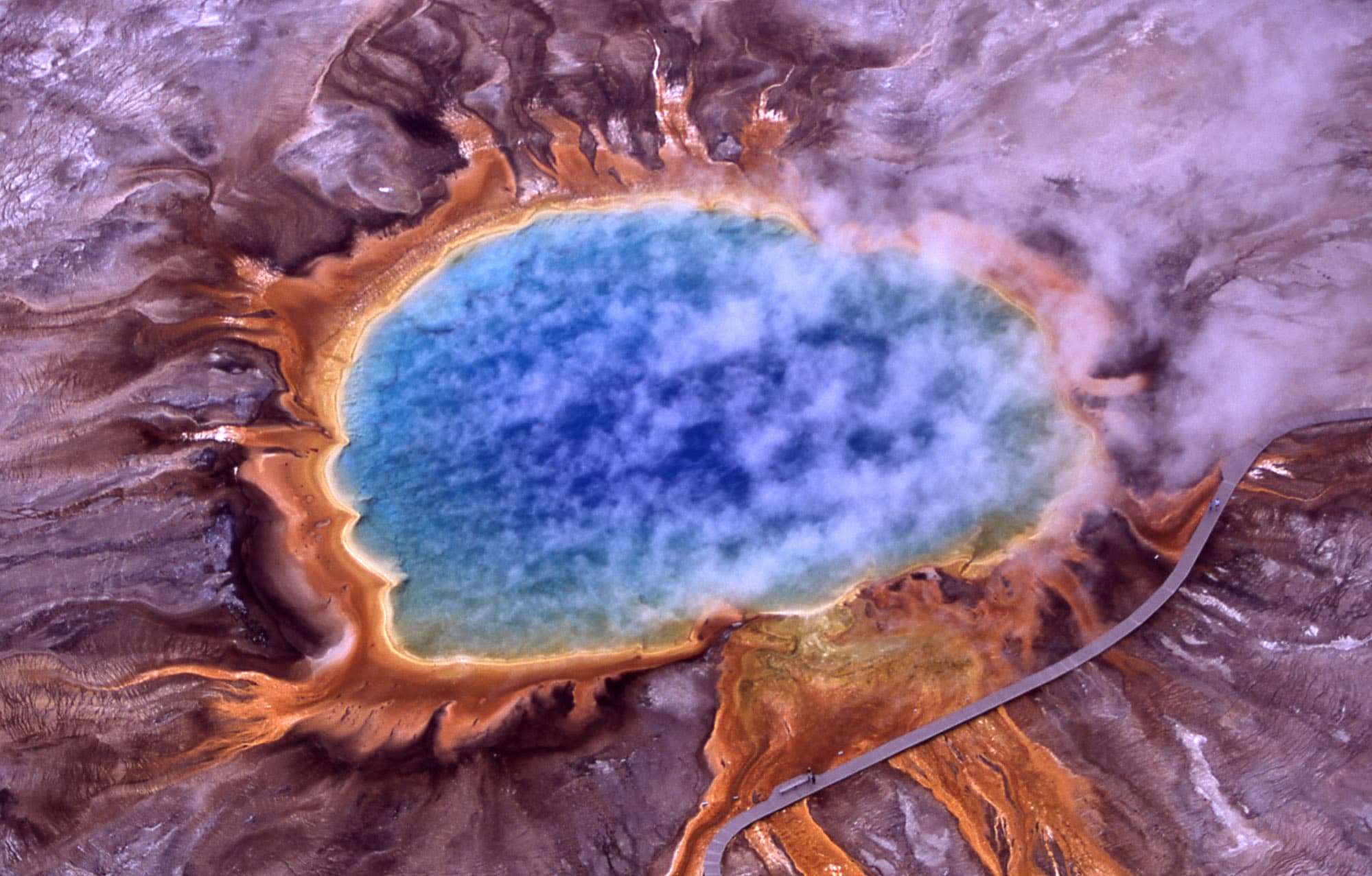 Vue aérienne d'une grande source chaude très colorée dans le parc national de Yellowstone, avec des anneaux orange, jaune, vert et bleu autour d'un centre bleu profond.