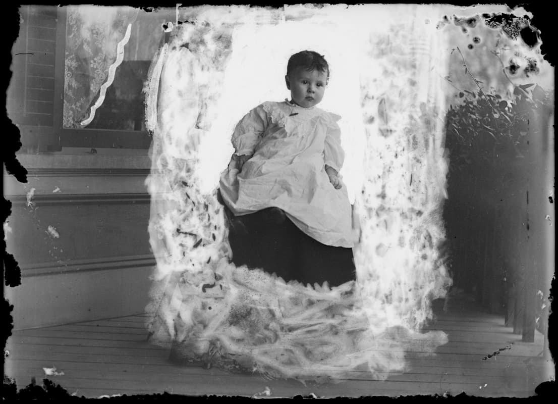 Photographie ancienne en noir et blanc d'un bébé de huit mois assis sur les genoux d'un adulte.