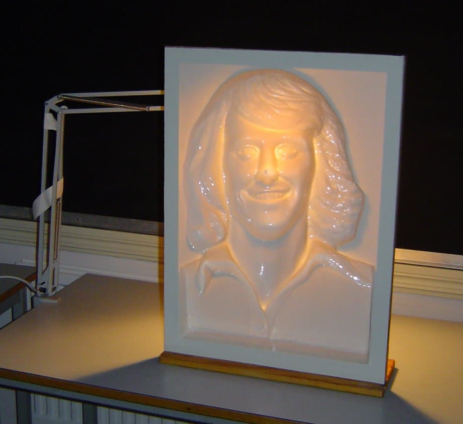 Un masque blanc de visage souriant monté dans un cadre et éclairé par une lampe de bureau ; il semble bomber vers l'extérieur.