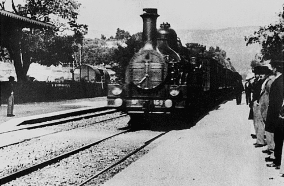 Image en noir et blanc d'une locomotive à vapeur arrivant le long d'un quai de gare, vue en biais, avec des voyageurs qui attendent.