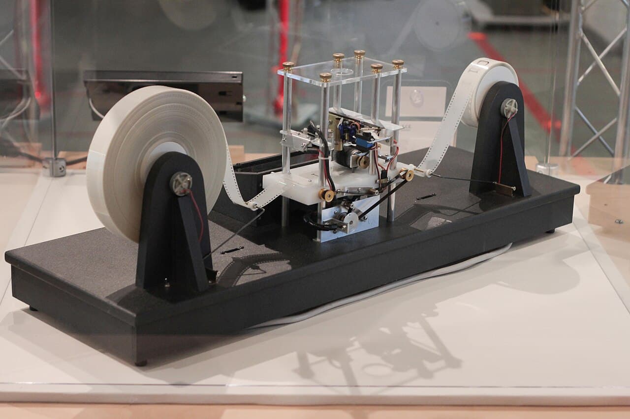 A physical model of a Turing machine: a long tape wound between two reels, passing under a read-write head on a wooden base.