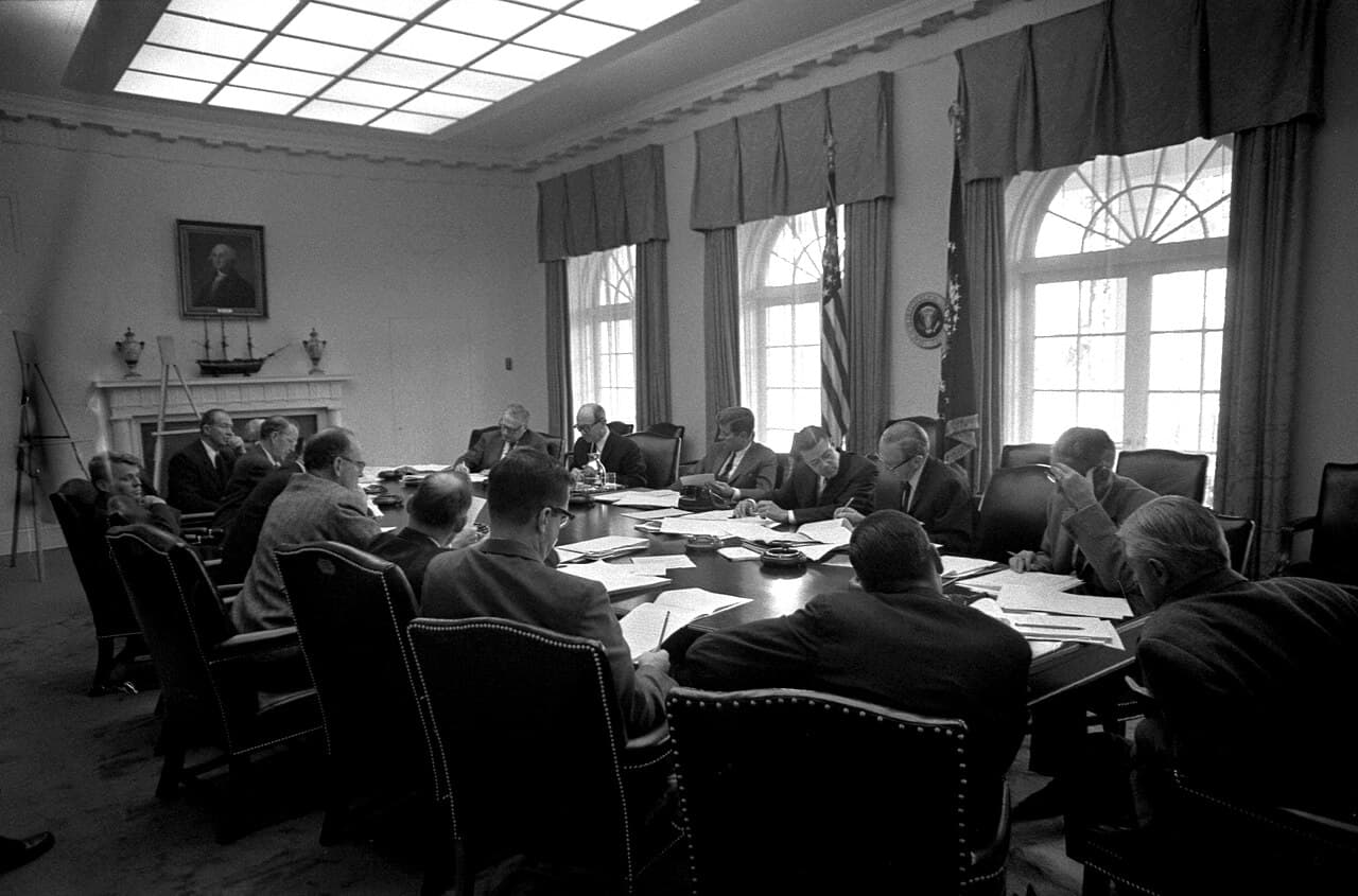 Photographie en noir et blanc du président Kennedy et de ses conseillers assis autour d'une table lors d'une réunion de l'EXCOMM à la Maison-Blanche.