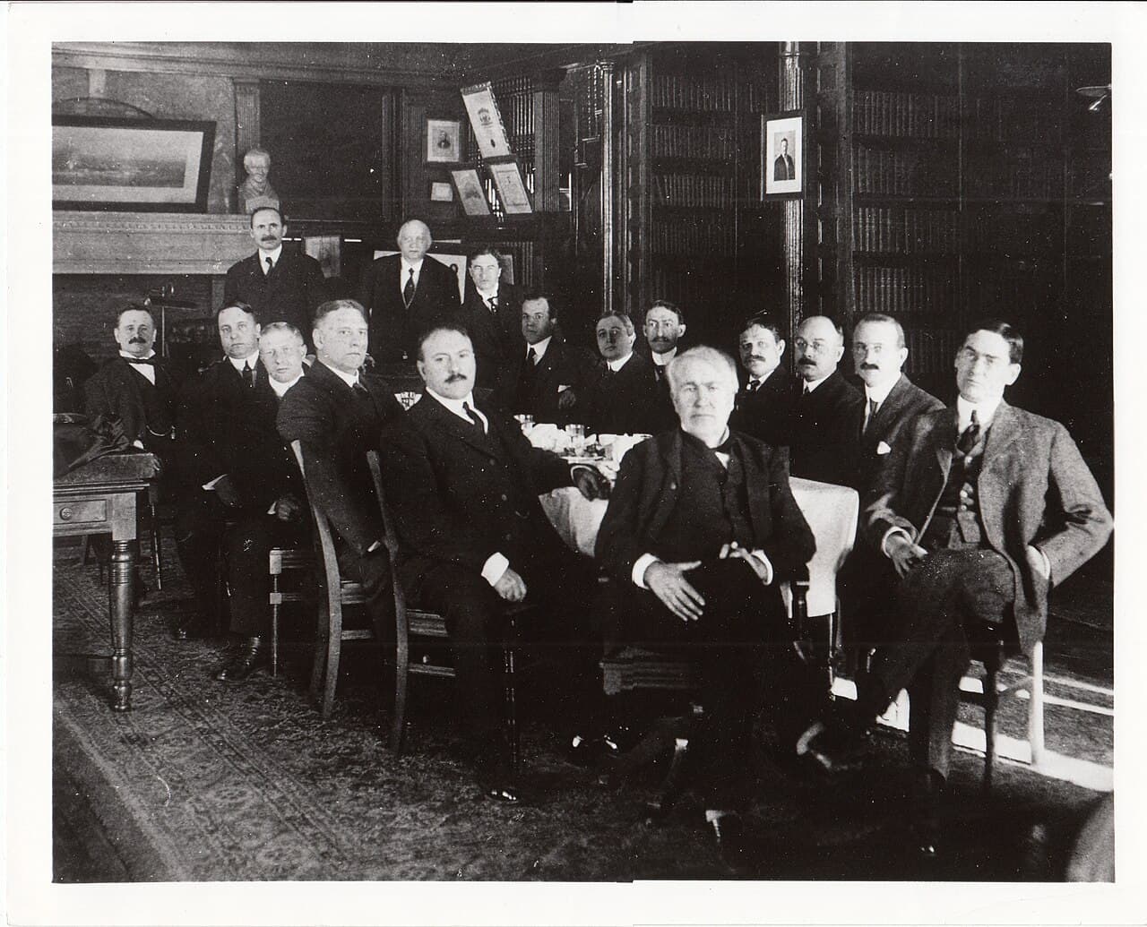 Un groupe d'hommes en costume sombre posant à l'intérieur autour de Thomas Edison, sur une photo en noir et blanc.