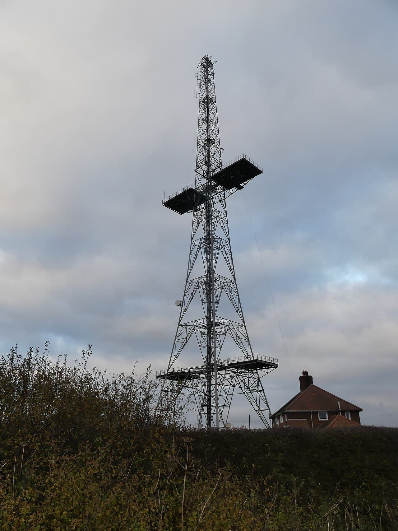 Une haute tour radio en treillis d'acier dressée dans un champ verdoyant, un mât émetteur survivant du réseau radar Chain Home.