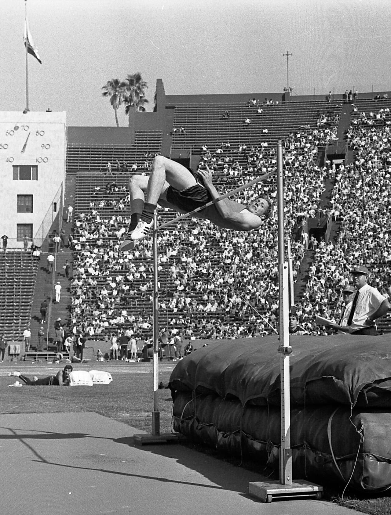 Photo en noir et blanc de Dick Fosbury cambré sur le dos au-dessus d'une barre de saut en hauteur, tête et épaules déjà passées