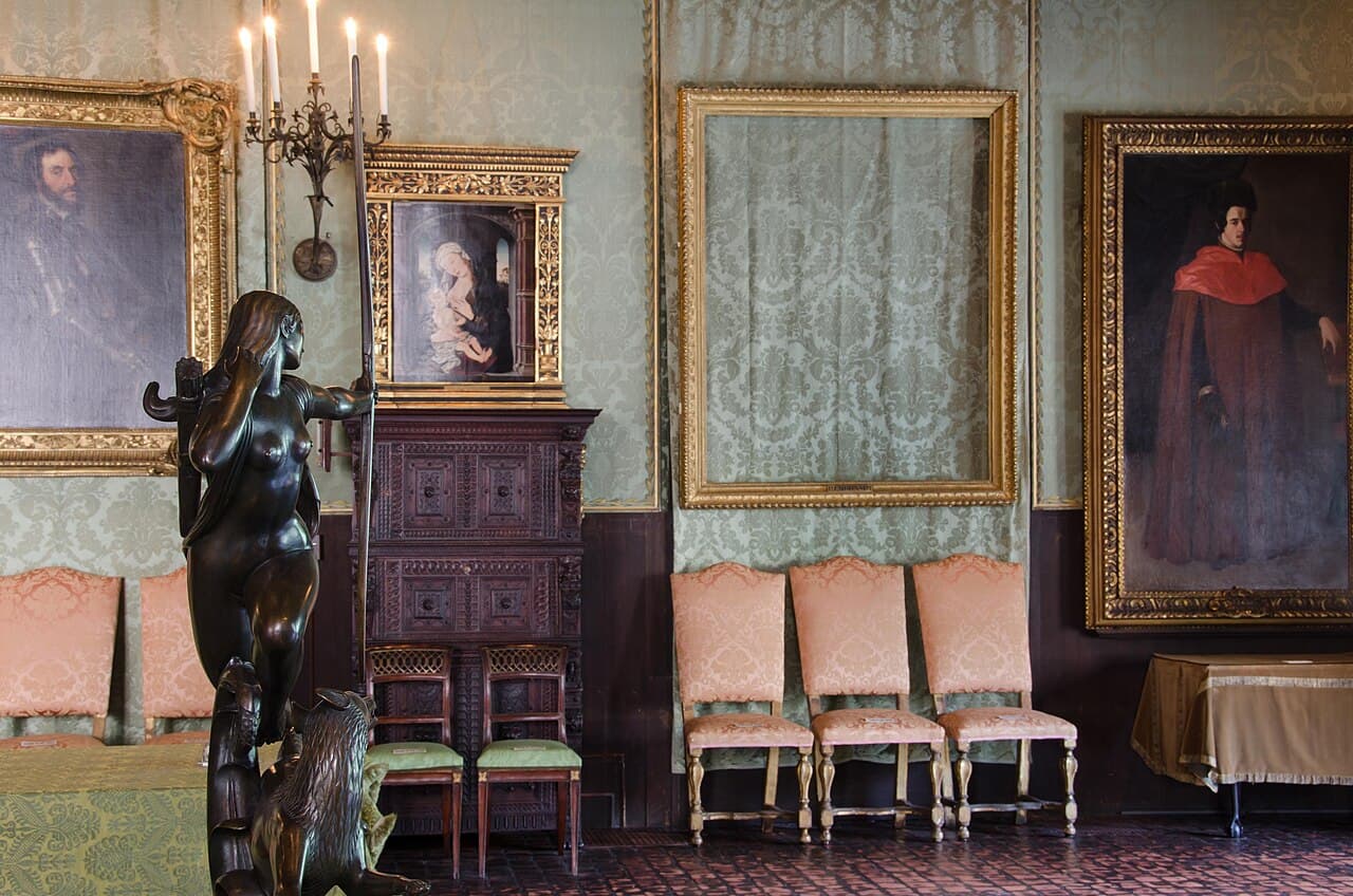 A museum room with green silk walls and gilded frames; in the middle of the wall, a large ornate gold frame hangs empty above a row of chairs.