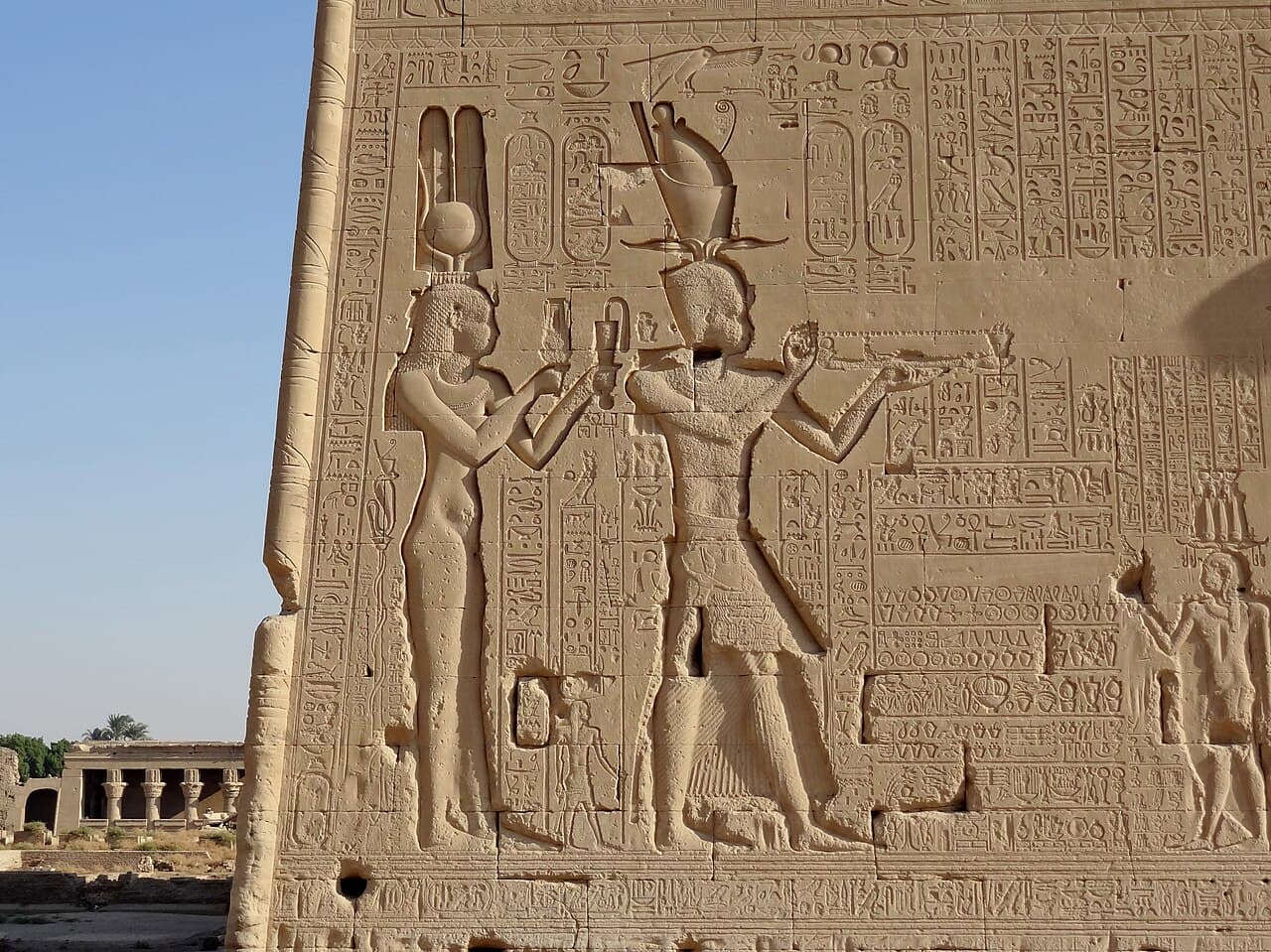 Un grand relief sculpté sur un mur de temple montrant deux personnages de profil, coiffés de couronnes royales égyptiennes, qui présentent des offrandes à des dieux égyptiens.