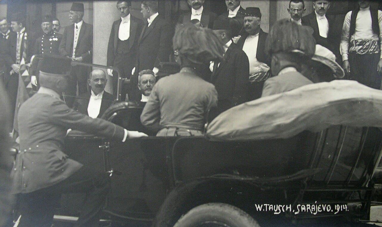 Black-and-white photo of an open-top car with a uniformed man and a woman in a white hat, surrounded by officials outside a building.