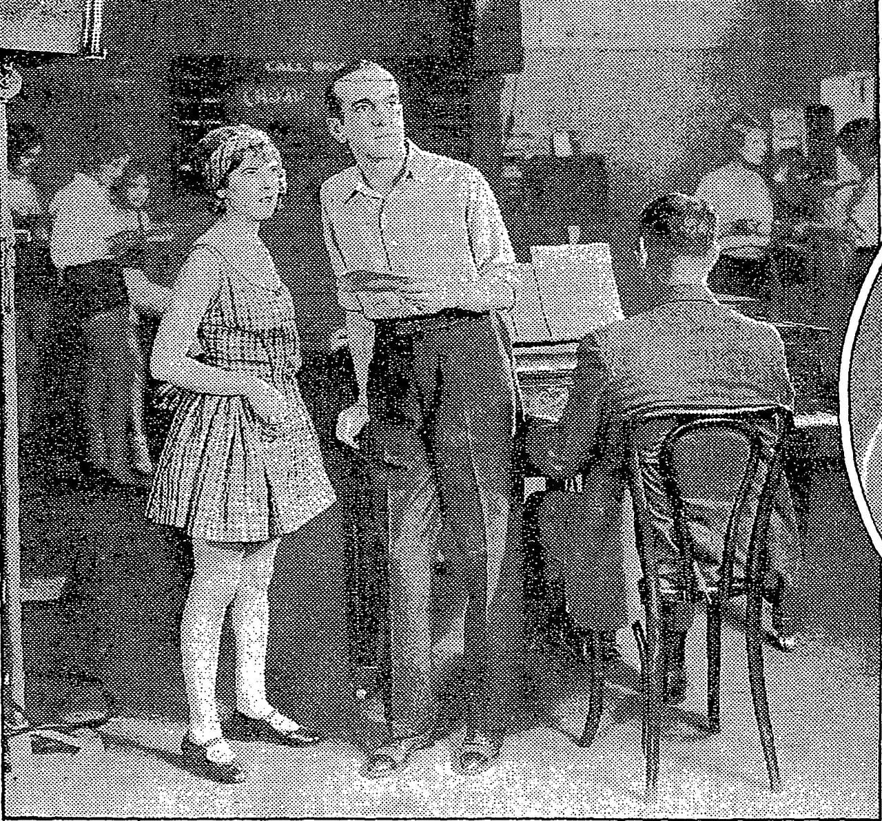 Black-and-white press photo of Al Jolson and May McAvoy standing by a piano on a stage set, with a pianist seated beside them.